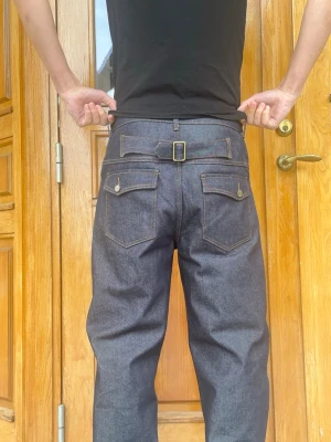 Raw Denim Jeans - Säljer ett par mörkblå loose fit jeans i rådenim med coolt spänne bak i midjan och kontrastsömmar. Jeansen har raka ben, två bakfickor med lock och knapp samt klassiska framfickor. Perfekta för dig som gillar en avslappnad och trendig stil.