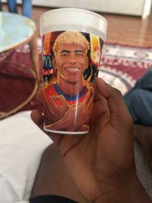 Plastmugg med fotbollstryck - Cool plastmugg med färgglatt tryck av en fotbollsspelare, eldflammor och texten 'LAMINE YAMAL'. Perfekt för dig som gillar sport och vill ha en unik mugg till drycken. Muggen är vit med detaljer i rött, orange och blått.