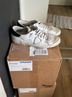 Vita låga sneakers från Philippe Model - Snygga vita sneakers från Philippe Model med klassisk låg siluett. Skorna har svarta detaljer på hälen, vita skosnören och en ikonisk sköld på sidan. Tillverkade i läder med slitstark gummisula. Perfekta för en clean och stilren look.