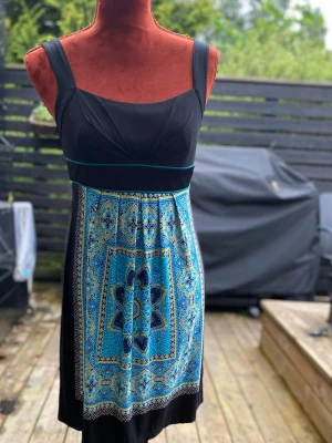 Svart och blå mönstrad kort klänning Strl: 6 - Klänning med en svart överdel och en mönstrad kjoldel i blått, turkos och gult. Klänningen har breda axelband och en markerad midja med ett turkost band. Baktill finns ett knytband i midjan. Baddräktsklänning L: 85 cm Bystvidd: 38 cm 95% polyester 5  % elastan  Inga bilder på mig! Katter finns i hemmet!