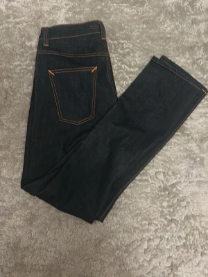 Mörkblå jeans från Nudie Jeans - Säljer ett par nya nudies använda en gång, mörkblå jeans från Nudie Jeans med raka ben och klassiska kontrastsömmar i orange. Jeansen har två bakfickor och en brun läderpatch med logga bak i midjan. Materialet är kraftig denim som ger en snygg och tidlös look.