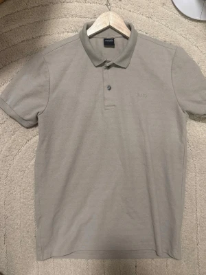 Beige pikétröja från BOSS - Klassisk beige pikétröja från BOSS med krage och två knappar framtill. Tröjan har korta ärmar och diskret BOSS-logga broderad på bröstet. Perfekt för en clean och stilren look.