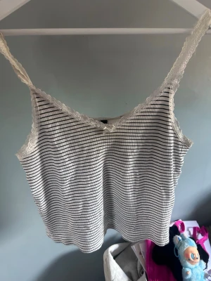 Randig vit linnetopp med spets - Söt vit linnetopp med tunna svarta ränder och spetsdetaljer längs axelbanden och ringningen. Toppen har en liten rosett framtill och är tillverkad i ett mjukt, ribbat material. Perfekt för varma dagar eller som layering-piece.