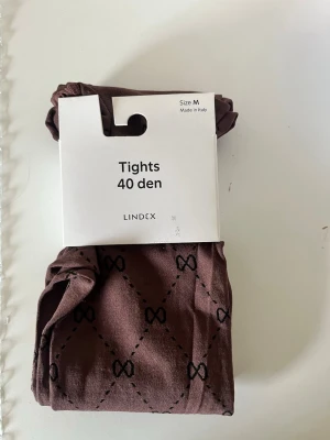 Bruna tights med mönster från Lindex - Bruna tights från Lindex, 40 den, med svart diskret rutmönster och små rosetter. Tillverkade i Italien. Perfekta för att ge din outfit en extra touch med sitt snygga mönster och tunna material.