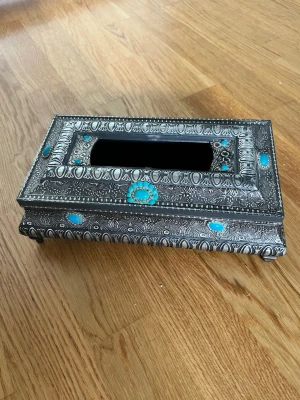 Dekorativ silverfärgad servetthållare - Säljer en rektangulär servetthållare i metall med detaljerade orientaliska mönster och turkosa stenar. Locket är avtagbart och har ett stort öppet fack för servetter. Perfekt som snygg inredningsdetalj på bordet. Oanvänd lite vintage stil 