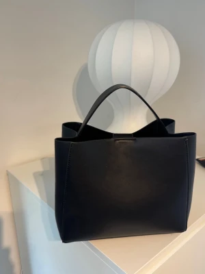 Svart stilren handväska i skinnimitation GINATRICOT - Snygg svart handväska i skinnimitation med minimalistisk design och raka linjer. Väskan har två handtag, magnetlås och ett innerfack med dragkedja. Perfekt för dig som gillar enkel och modern stil.