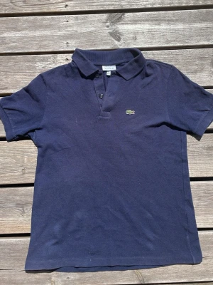 Marinblå pikétröja från Lacoste - Klassisk marinblå pikétröja från Lacoste med korta ärmar och krage. Tröjan har två knappar framtill och den ikoniska gröna krokodilloggan broderad på bröstet. Tillverkad i mjuk bomull som känns skön mot huden. Passar till allt. Perfekt inför sommaren. Najs att golfa i