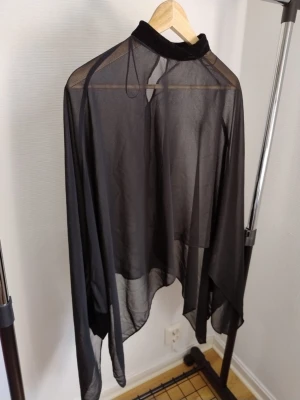 Svart transparent blus med polokrage - Stilren svart blus i transparent material med hög polokrage. Blusen har långa ärmar och är tillverkad i ett tunt, lätt tyg som ger en luftig känsla. Perfekt för att skapa en trendig lager-på-lager-look.