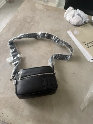 Svart axelväska från Karl Lagerfeld - Snygg svart axelväska från Karl Lagerfeld med justerbar axelrem och silverfärgad dragkedja. Väskan har en stilren, rektangulär form och ett diskret KL-logotypmärke på framsidan. Tillverkad i slätt konstläder som ger en modern och clean look.
