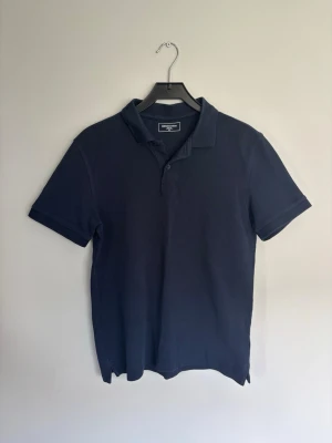 Mörkblå pikétröja från Dressmann - Klassisk mörkblå pikétröja från Dressmann med korta ärmar och krage. Tröjan har knappar vid halsen och är tillverkad i ett mjukt bomullsmaterial som känns skönt mot huden. Perfekt till jeans eller shorts för en enkel och stilren look.