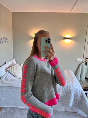 From Future tröja  - Cool grå stickad tröja med neonrosa ribbade muddar och halsringning från From Future. Text i rosa längs båda ärmarna. Tröjan är lite kortare i modellen och har en avslappnad passform. 