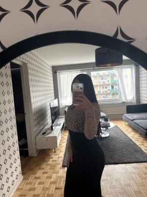 Svart långklänning med glittrig topp - HELT NY LAPPEN ÄR KVAR! Elegant svart långklänning med glittrig, silverfärgad topp och långärmade meshärmar. Klänningen har offshoulder-design och figurnära passform, perfekt för speciella tillfällen. Materialet ser ut att vara polyester med inslag av glitterdetaljer.