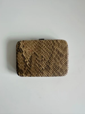 Brun plånbok / korthållare i ormmönster - Stilren Korthållare / plånbok med ormskinnsmönster. Insidan har fack för kort och ett genomskinligt fack. Stängs med ett metallspänne. 9x12cm. Mycket gott skick!