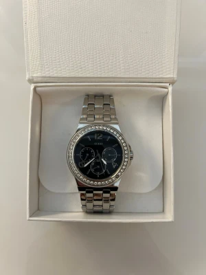 Silverfärgad klocka från Guess - Stilren klocka från Guess i silverfärgat metallarmband och svart urtavla. Urtavlan har tre mindre visare och är dekorerad med gnistrande stenar runt boetten. Perfekt accessoar för dig som gillar en elegant look. OBS! (En sten saknas, men det är inget som påverkar helhetsintrycket).
