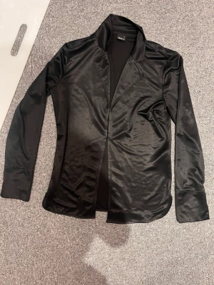 Svart satinliknande kavaj - Stilren svart kavaj i ett glansigt, satinliknande material. Kavajen har klassisk krage, två knappar framtill och långa ärmar. Perfekt för dig som vill ha en elegant och trendig look.