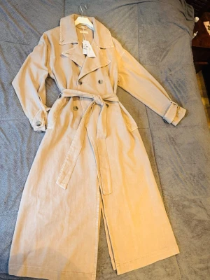 Beige jeans kapp från Ginatricot - Stilren beige trenchcoat från GinaTricot i mjuk manchester. Dubbelknäppt med bred krage, knytskärp i midjan och fickor på sidorna. Långa ärmar med justerbara remmar vid ärmslut. Perfekt för dig som gillar klassisk men trendig look.