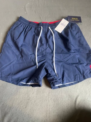 Marinblå badbyxor Polo Ralph Lauren - Snygga marinblå badbyxor från Polo Ralph Lauren med vit dragsko i midjan och röd detalj på insidan av linningen. Klassisk modell med meshfoder och diskret logga på benet. Perfekta för stranden eller poolen.