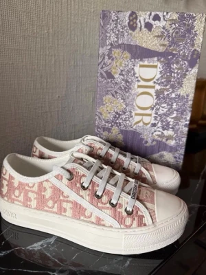 walk n dior platform sneaker - Säljer ett par snygga Dior sneakers i rosa och vitt med det klassiska Dior-mönstret. Skorna har vita snören, gummisula och detaljer med 'Christian Dior' på tungan. Perfekta för dig som vill ha en exklusiv och trendig look.