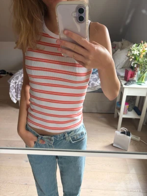 Randig ribbad linnetopp - Säljer en vit ribbad linnetopp med orangea horisontella ränder. Toppen är ärmlös och har en tight passform med rund halsringning. Perfekt till jeans för en avslappnad look.