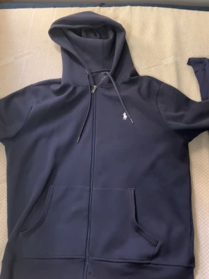 Mörkblå hoodie från Polo Ralph Lauren - Mörkblå hoodie från Polo Ralph Lauren med dragkedja framtill, justerbar huva med snören och klassisk vit logga på bröstet. Tillverkad i mjukt bomullsmaterial och har en stor känguruficka framtill. Perfekt för en avslappnad och stilren look.