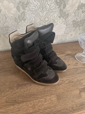 Svarta Isabel Marant bekett sneakers - Säljer ett par svarta bekett sneakers från Isabel Marant med högt skaft och breda kardborreband. Skorna har en chunky sula och är gjorda i mocka och läder med beige insida. Perfekta för dig som vill ha en edgy och trendig look. Passar även dig som har 39