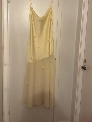 Ljusgul långklänning med tunna band - Elegant ljusgul långklänning med tunna axelband och diskreta blommiga detaljer. Klänningen har en mjuk, flytande siluett och är tillverkad i ett glansigt material som ger en lyxig känsla. Perfekt för sommarens alla tillfällen.