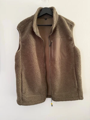 Brun teddyväst med dragkedja - Mysig brun teddyväst med hög krage och hel dragkedja framtill. Västen har en bröstficka i nylon med dragkedja och är tillverkad i mjukt, fluffigt material. Perfekt för lager på lager och ger en avslappnad look.