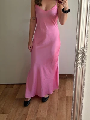 Rosa långklänning i satin - Snygg rosa långklänning med smala axelband och figurnära passform. Klänningen har en slät, glansig yta som ger en lyxig känsla och faller fint längs kroppen. Perfekt för sommarens alla tillfällen.