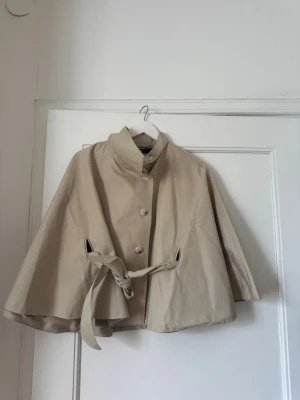 Beige cape trenchcoat med knytband - Stilren beige trenchcoat i cape-modell med knytband i midjan och knappar framtill. 