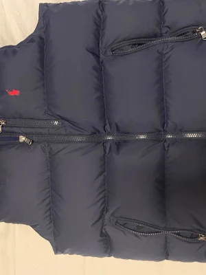 Mörkblå dunväst från Polo Ralph Lauren - Snygg mörkblå dunväst från Polo Ralph Lauren med klassisk röd logga på bröstet. Västen har flera dragkedjeförsedda fickor och quiltad design. Perfekt för lager på lager och håller dig varm under kyliga dagar.