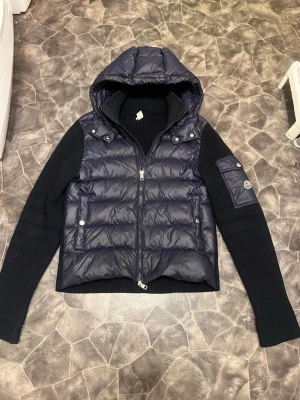 Mörkblå pufferjacka från Moncler - Snygg mörkblå pufferjacka från Moncler med stickade svarta ärmar och huva. Jackan har två fickor med tryckknappar, dragkedja framtill och en Moncler-logga på ärmen. Perfekt för kalla dagar och riktigt trendig look, han på bilden är runt 170cm! 65kg, skriv vid intresse. Kom gärna med bud! 🙌 