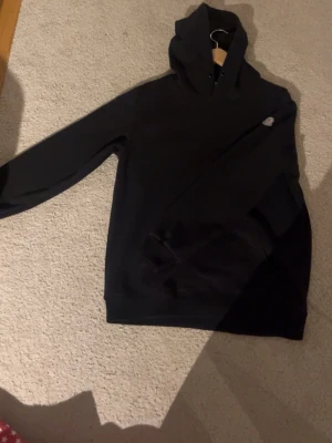 Svart hoodie från Moncler - Säljer en svart hoodie från Moncler med klassisk huva och lång ärm. Hoodien har en liten Moncler-logga på ärmen och är tillverkad i mjukt bomullsmaterial. Perfekt för dig som gillar stilrena och exklusiva plagg.
