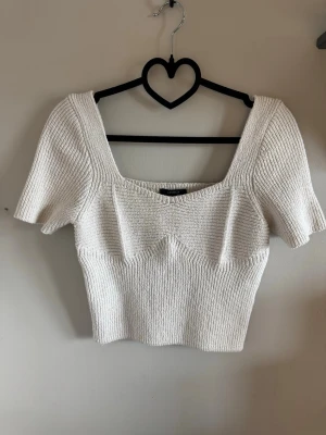 Ljusbeige ribbad stickad croppad topp - Trendig ljusbeige croppad topp med ribbad stickning och fyrkantig ringning. Toppen har korta ärmar och en figurnära passform som framhäver siluetten. Perfekt att styla med höga jeans eller kjol för en cool look.