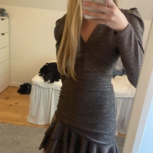 Glittrig klänning - Säljer en snygg glittrig kort klänning med v-ringning och långa puffärmar. Klänningen är croppad med en volang längst ned. Perfekt när man ska vara lite extra fin. 