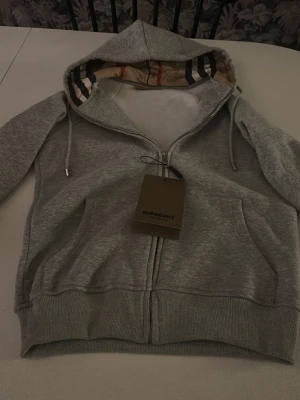 Grå hoodie från Burberry med dragkedja Stl 152 - Säljer en grå hoodie från Burberry med klassiskt rutigt foder i huvan och dragkedja framtill. Hoodien har ribbade muddar, fickor och snören i huvan. Materialet är mjukt och bekvämt, perfekt för en avslappnad look. Helt ny med tag 