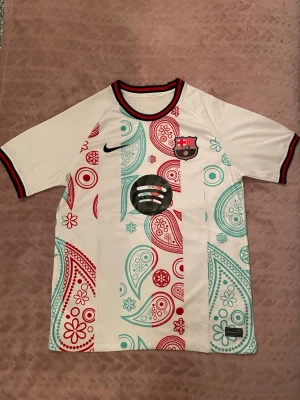 Barcelona paisleymönstrad matchtröja - Säljer en unik FC Barcelona matchtröja från Nike med paisleymönster i rött och turkost på vit botten. Tröjan har klubbmärke, Nike-logga och Spotify-logga på bröstet samt svarta och röda detaljer vid krage och ärmslut. Perfekt för dig som älskar fotboll och snygga matchtröjor.