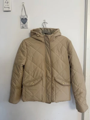 Beige pufferjacka från U.S. Polo Assn. - Snygg beige pufferjacka från U.S. Polo Assn. med quiltat mönster, huva med dragsko och två fickor med guldfärgade knappar. Jackan har dragkedja och tryckknappar framtill samt diskret logga broderad på bröstet. Perfekt för kalla vinterdagar.