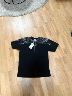 Svart t-shirt Marcelo Burlon vingar - Svart t-shirt från Marcelo Burlon County of Milan med ikoniskt vingtryck i vitt och grått över axlar och bröst. Klassisk passform och rund hals. Perfekt för dig som vill sticka ut med en streetwear vibe.