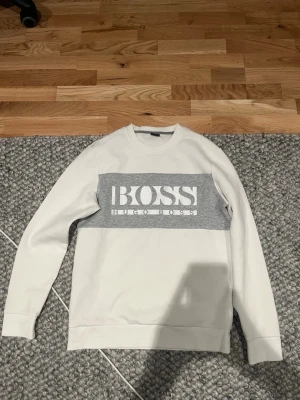 Vit sweatshirt från Hugo Boss - Snygg vit sweatshirt från Hugo Boss med grå panel och stor vit logga på bröstet. Tröjan har rund halsringning, långa ärmar och ribbade muddar. Perfekt för en clean och stilren look.