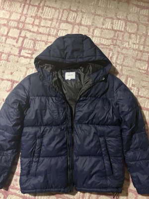 Mörkblå pufferjacka från Jack & Jones - Säljer en mörkblå pufferjacka från Jack & Jones med huva och dragkedja framtill. Jackan har två sidofickor och quiltad design som ger en schysst streetstil. Perfekt för kalla dagar och riktigt skön att bära. Materialet är syntet och jackan är fodrad för extra värme.