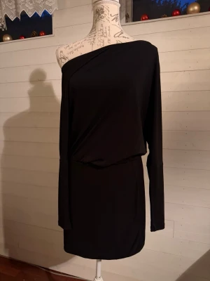 Svart oneshoulder kort klänning - Snygg svart kort klänning med oneshoulder-design och lång ärm på ena sidan. Klänningen har en lösare passform upptill och smalare kjol, vilket ger en cool och modern siluett. Tillverkad i mjukt, stretchigt material som känns bekvämt mot huden.