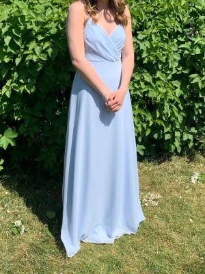 Ljusblå långklänning med spetsdetaljer - Elegant ljusblå balklänning med omlott framtill och tunna axelband. Klänningen har v-ringning och vacker spets i ryggen 