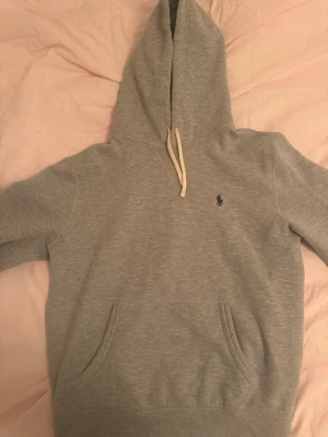 Grå hoodie från Polo Ralph Lauren - Snygg grå hoodie från Polo Ralph Lauren med klassisk huva och vita dragsnören. Broderad marinblå logga på bröstet och stor magficka framtill. Tillverkad i mjuk bomullsblandning som känns skön mot huden. Perfekt för en avslappnad och stilren look.
