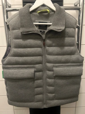 Grå quiltad väst från Elias Rumelis - Snygg grå väst från Elias Rumelis med quiltad design och bred ribbad krage. Västen har två stora fickor framtill och stängs med dragkedja. Perfekt för våren som kommer! 