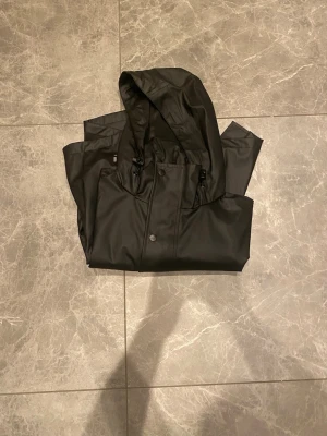 Svart regnjacka från Helly Hansen - Snygg svart regnjacka från Helly Hansen med huva och tryckknappar framtill. Jackan är lång i modellen och har justerbar dragsko vid huvan. Perfekt för regniga dagar och enkel att matcha med olika outfits.