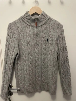 Grå kabelstickad tröja Polo Ralph Lauren - Kabelstickad grå tröja från Polo Ralph Lauren med halv dragkedja och hög krage. Tröjan har ribbade muddar och nederkant samt den klassiska broderade loggan på bröstet. Perfekt för lager på lager och riktigt snygg till jeans.