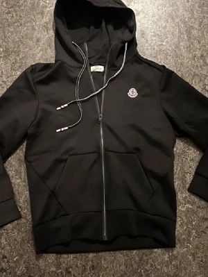 Svart Moncler hoodie med dragkedja - Snygg svart hoodie från Moncler med dragkedja framtill och klassisk Moncler-logga på bröstet. Huva med dragsko och detaljer i rött, vitt och blått på snörena. Tillverkad i mjukt och skönt material. Perfekt för en avslappnad och trendig look.