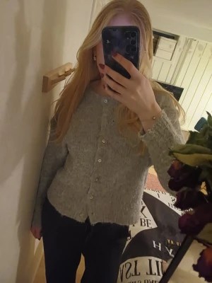 Grå stickad kofta med pärlemorknappar - Mysig grå stickad kofta med rund hals och pärlemorskimrande knappar framtill. Koftan har lång ärm och en lätt fluffig struktur som ger en avslappnad look. Perfekt att slänga över en topp för extra värme.