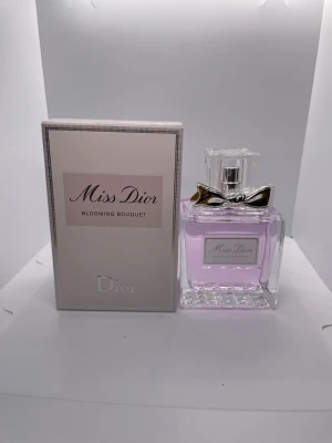 Miss Dior Blooming Bouquet parfym - Miss Dior Blooming Bouquet från Dior är en elegant parfym med ljusrosa doftvätska i en stilren, fyrkantig glasflaska. Flaskan har en silverfärgad rosett vid halsen och ett genomskinligt lock. Förpackningen är ljus med subtila rosa och silverdetaljer.