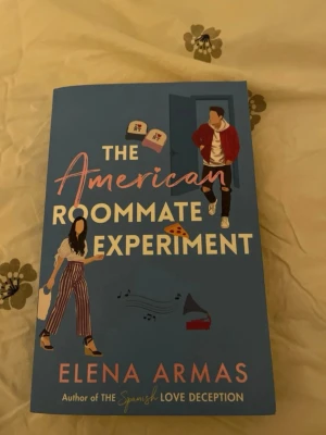 The American Roommate Experiment - En romantisk komediroman på engelska av Elena Armas. Omslaget är blått med illustrationer av två personer, pizza, böcker och en grammofon. Boken handlar om Rosie och Lucas som delar lägenhet i New York och utforskar kärlek och kreativitet.